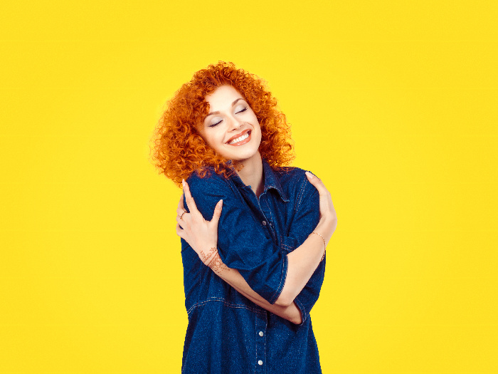 A woman hugging herself with a smile on her face against a yellow background