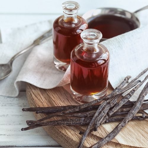 Two vanilla extract bottles next to fresh vanilla beans and a bowl of vanilla extract with a spoon