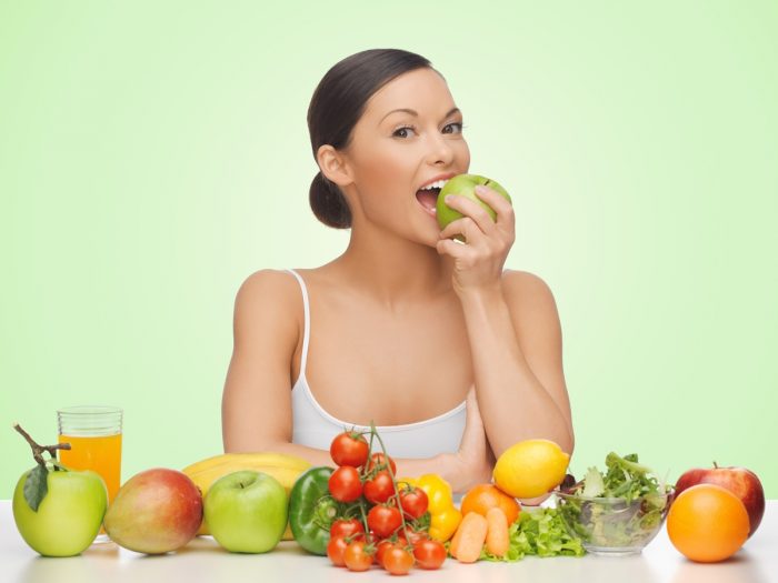 Woman biting an apple, sitting in front of an assortment of fruits.