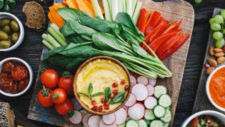 Board with cucumbers, tomatoes, red bells, celery, radishes, carrot sticks, spinach, and hummus