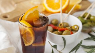 A glass of juice with slices of citrus fruit, ice cube, blurry images of bowl of olive and cut fruits at the back.