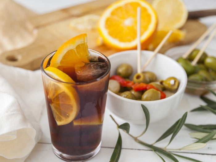 A glass of juice with slices of citrus fruit, ice cube, blurry images of bowl of olive and cut fruits at the back.