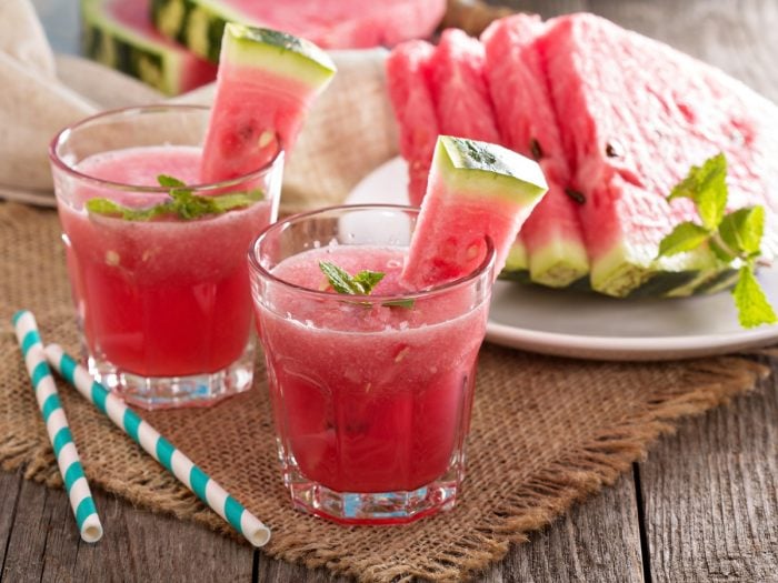 Two small glasses of watermelon juice accompanied by straws and watermelon slices on a table