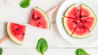 2 watermelon popsicles and a plate full of sliced watermelon on a white wooden background