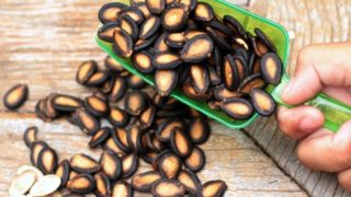 Hand holding a green plastic scoop with watermelon seeds over wooden background with more watermelon seeds
