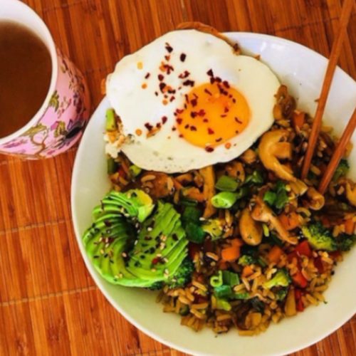 Stir-fried vegetables sauteed with rice, served with avocado and eggs to the side, placed atop a wooden table