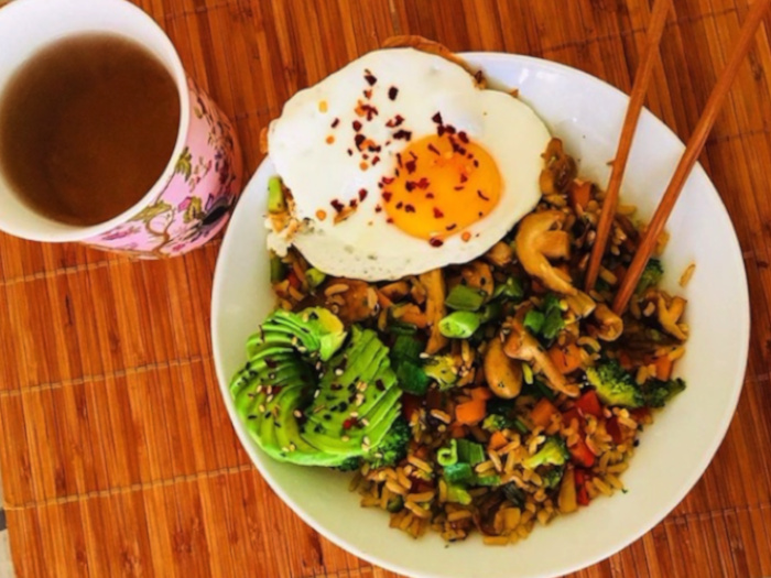 Stir-fried vegetables sauteed with rice, served with avocado and eggs to the side, placed atop a wooden table