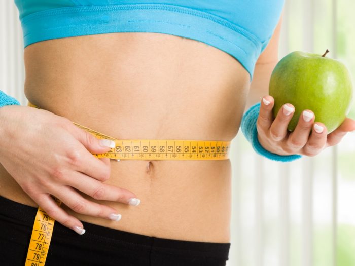 Woman measuring her waist with a measuring tape and holding an apple.