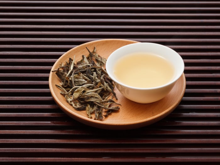 A white bowl of white tea placed on a wooden plate with dried white leaves