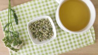 A flat lay picture of a cup of yarrow tea and a bowl of dried yarrow flowers with leaves on a wooden table