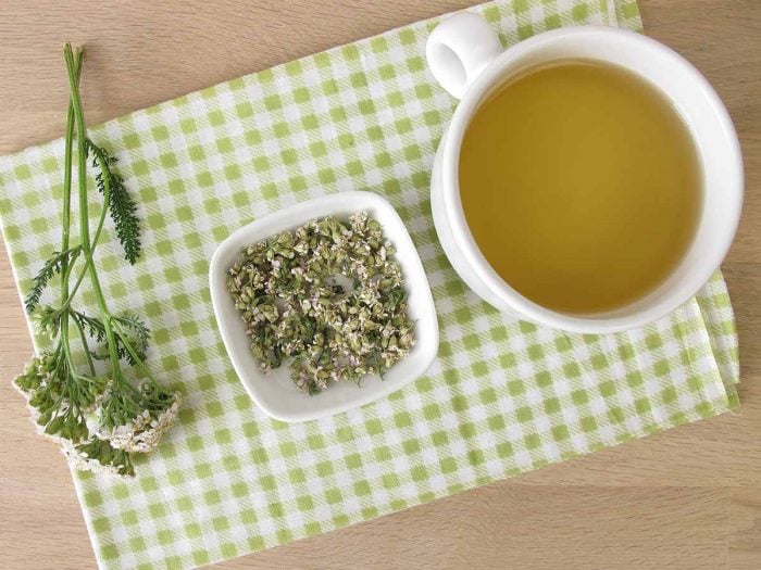A flat lay picture of a cup of yarrow tea and a bowl of dried yarrow flowers with leaves on a wooden table
