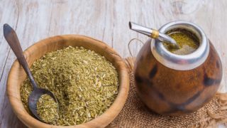 A wooden bowl of yerba mate and a jar of yerba mate tea with a metal straw on a wooden table