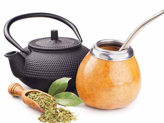 A tea pot and flask of yerba mate tea kept beside a spoon of dried yerba mate tea leaves against a white background