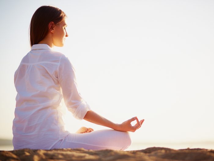 A young woman dressed in white clothes sitting in padmasana pose and practicing yoga on the beach