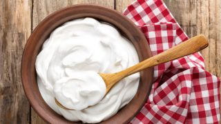 A wooden bowl filled with yogurt with a wooden spoon in it placed next to a red check cloth on a wooden table