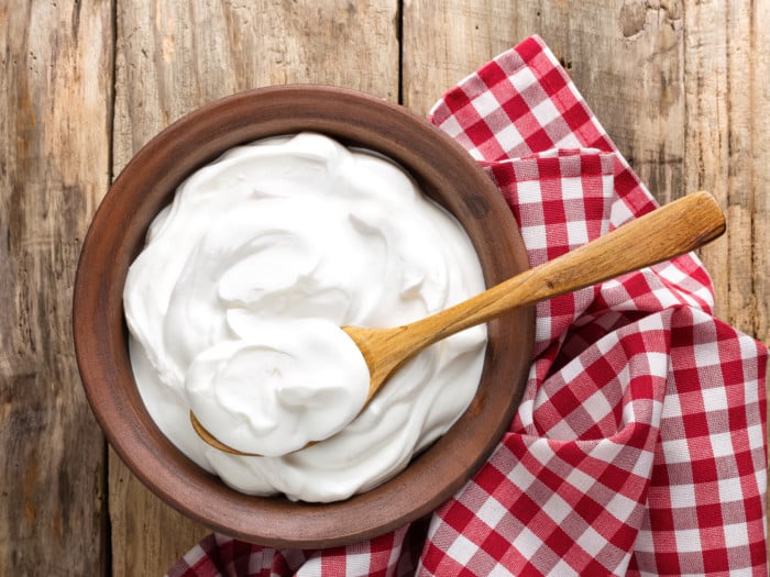 A wooden bowl filled with yogurt with a wooden spoon in it placed next to a red check cloth on a wooden table