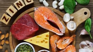 Foods rich in zinc (sliced salmon, meat, prawns, garlic, mushrooms, almonds, cashews, spinach, & legumes) on a wooden table