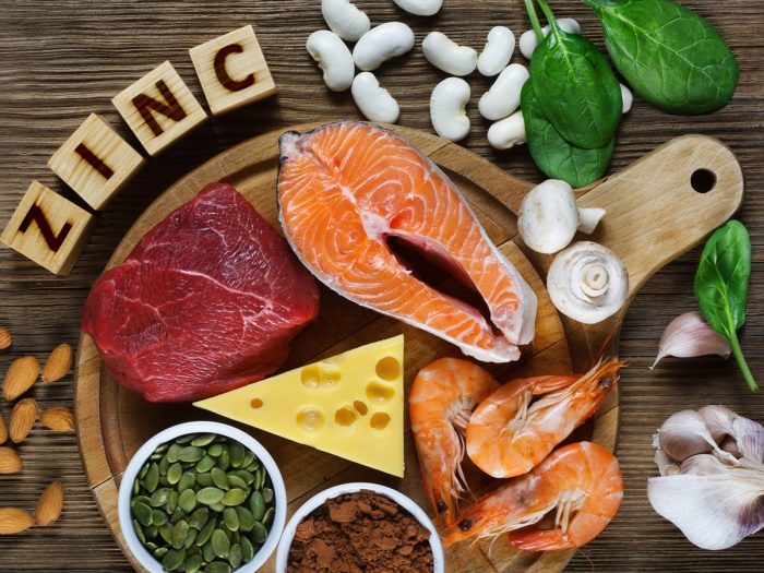 Foods rich in zinc (sliced salmon, meat, prawns, garlic, mushrooms, almonds, cashews, spinach, & legumes) on a wooden table